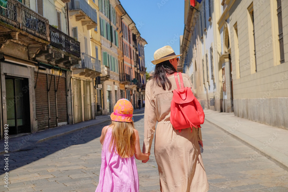Mother with a red backpack and a straw fedora holding hands with her young daughter in a pink dress and orange panama hat. tourist with a child walking down the European narrow street. sunny day