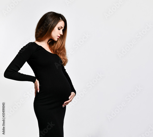 Brunette pregnant woman in black tight dress. She put hand on her belly while posing sideways isolated on white. Close up