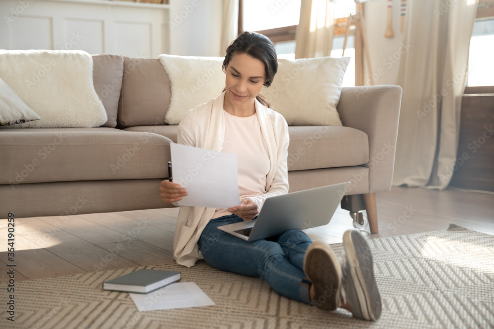 Young Caucasian woman sit on warm floor in living room working on laptop with paperwork documents, millennial girl relax at home, studying on modern computer gadget, online education concept Young Caucasian woman sit on warm floor in living room working on laptop with paperwork documents, millennial girl relax at home, studying on modern computer gadget, online education concept