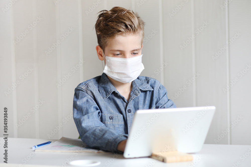 School boy with face mask watching online education classes at home. COVID-19 pandemic forces children online learning.