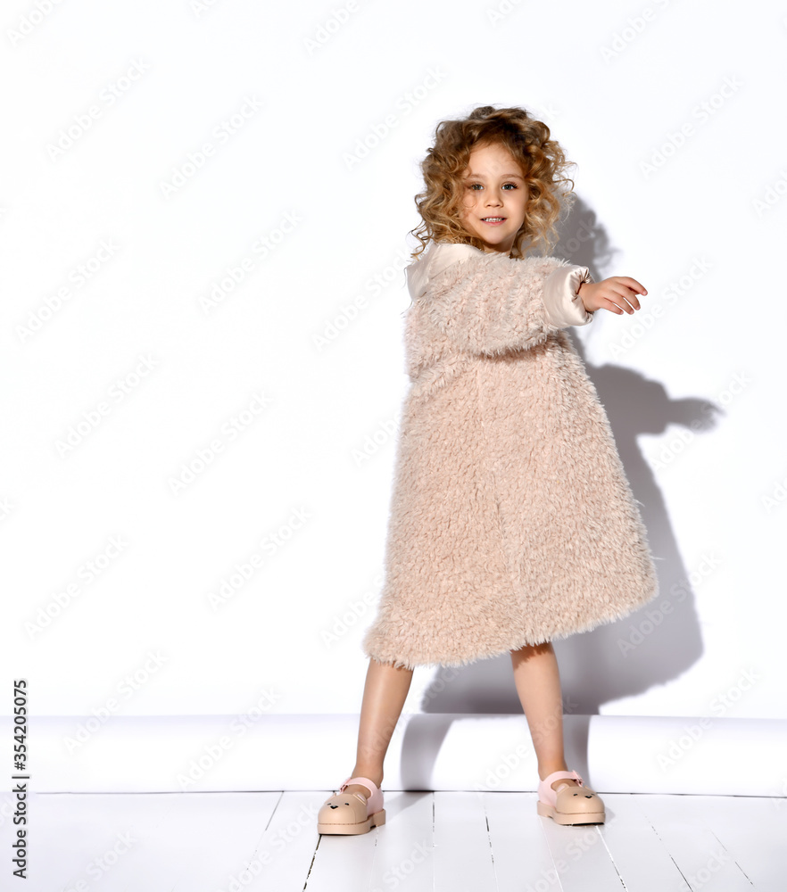 Little blonde model in golden necklace, fluffy pink dress and fur coat, shoes. Spread her hands, smiling, posing isolated on white Little blonde model in golden necklace, fluffy pink dress and fur coat, shoes. Spread her hands, smiling, posing isolated on white