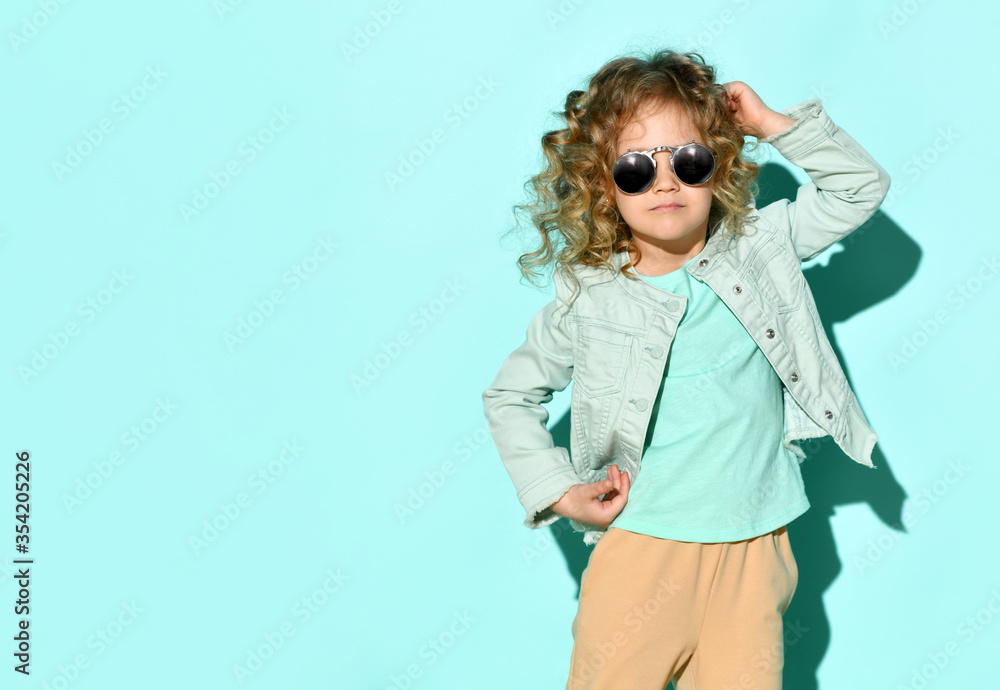 Adorable girl with curly hair in glasses with black lens looking up pensively holding one hand in pocket and touching neck by another one Adorable girl with curly hair in glasses with black lens looking up pensively holding one hand in pocket and touching neck by another one