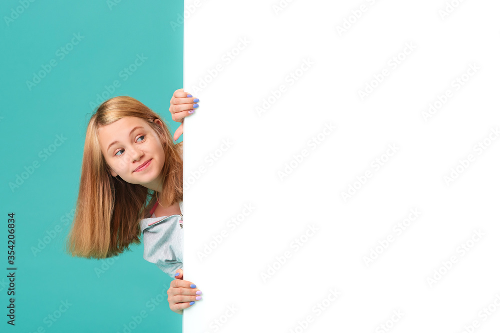 A smiling girl stands behind a white blank panel isolated against a turquoise background. Looks out from behind the banner, an empty space for the text. A smiling girl stands behind a white blank panel isolated against a turquoise background. Looks out from behind the banner, an empty space for the text.