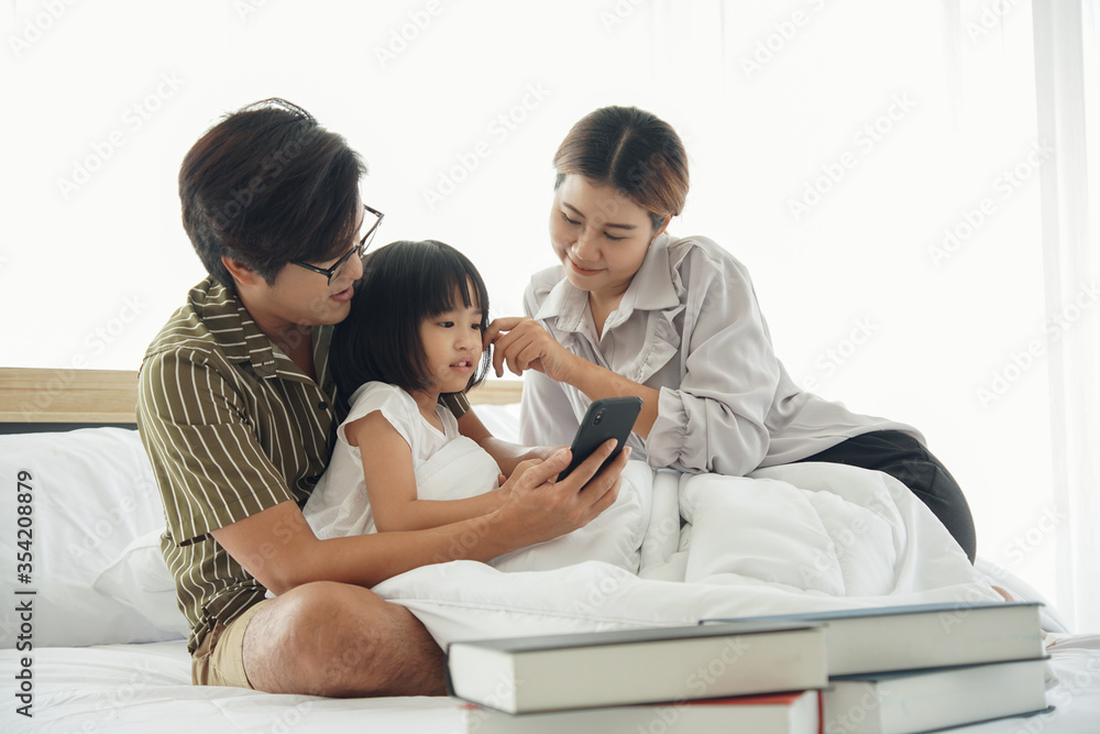 Happy asian young family spend time together. Dad hold mobile phone taking photo selfie with mom daughter who sitting under white blanket on the bed at bedroom