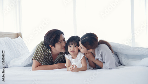 Asian Parents lying on bed under blanket and kissing their little daughter on both cheeks. family portrait