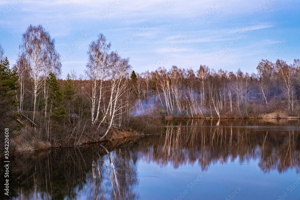 A steep shore overgrown with trees and bushes, the Uvod River and the blue smoke of a bonfire. A steep shore overgrown with trees and bushes, the Uvod River and the blue smoke of a bonfire.