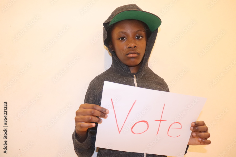 African American Child wearing hoodie and hat holding White paper sign with  word Vote written in red letters