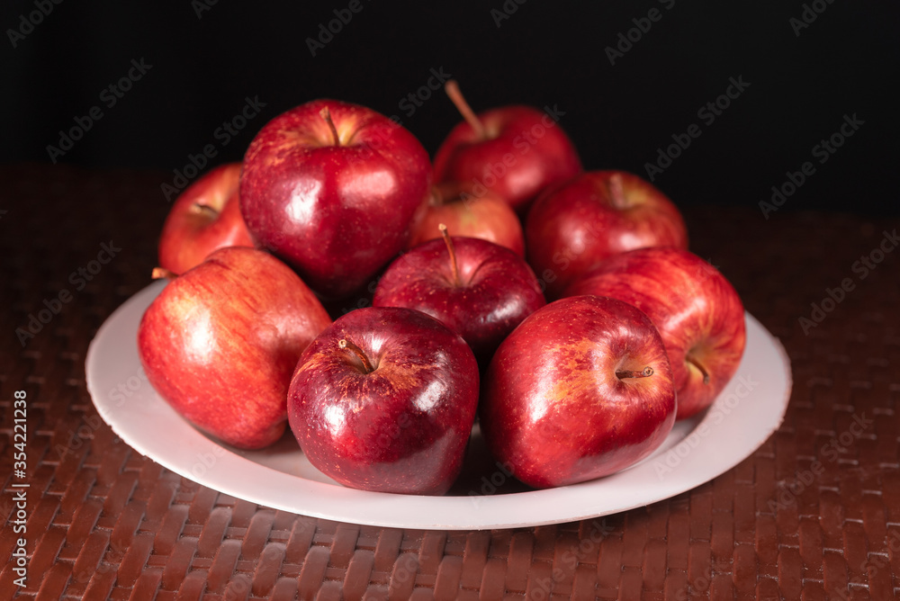 many red apples on white plate on brown woven table close-up and black background many red apples on white plate on brown woven table close-up and black background
