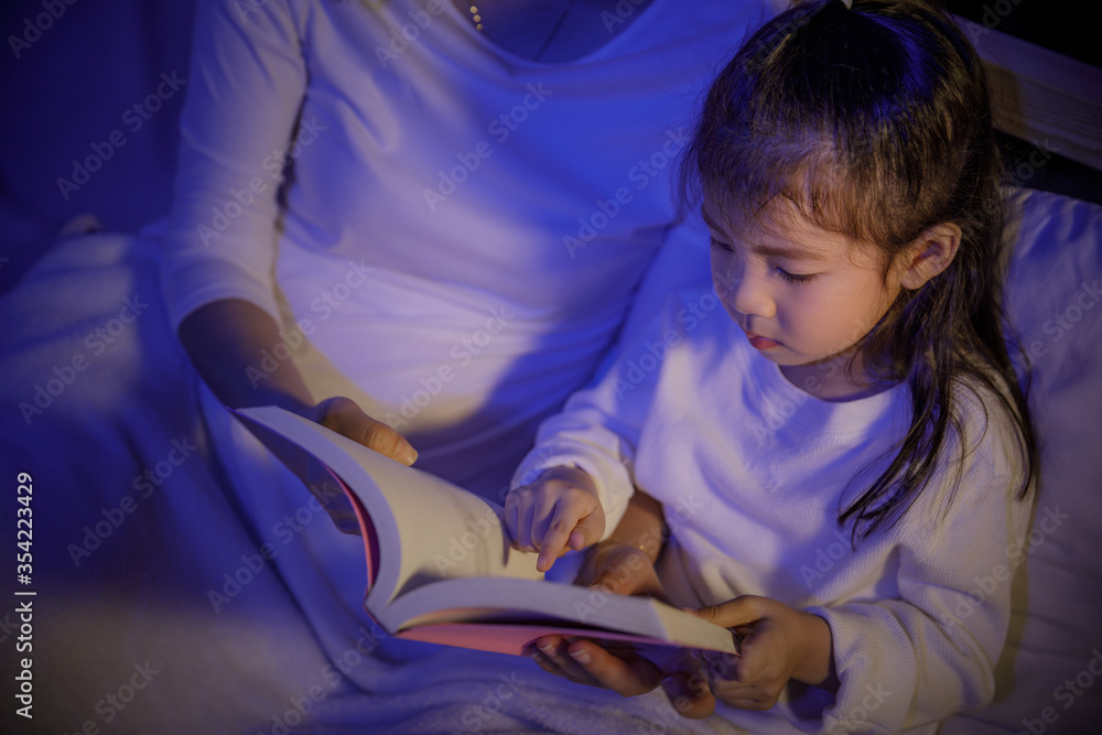 Mom reading a book with her daughter on bed in a dark bedroom at night before sleep, Parenthood and happy moments concept Mom reading a book with her daughter on bed in a dark bedroom at night before sleep, Parenthood and happy moments concept