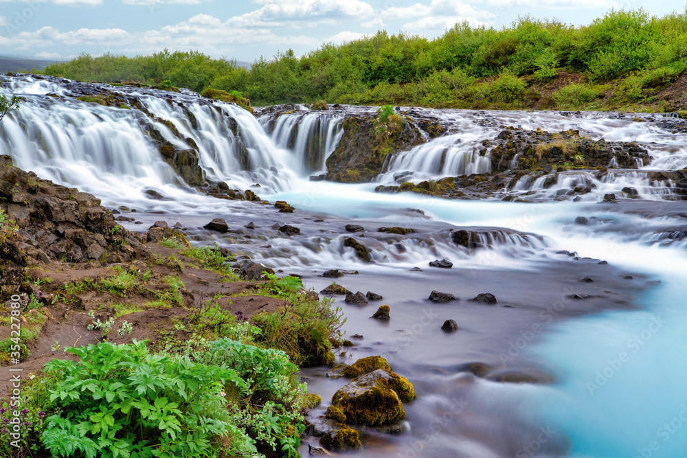 Bruarfoss Waterfall in the summer, with trees and green grass all over the area. This is a waterfall that is hidden among nature in Iceland.