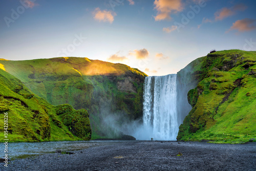 Fototapeta Naklejka Na Ścianę i Meble -  Men and women are standing and watch at the great Skogafoss Falls. In the morning, the sunrise comes from behind the mountains, with green grass all over the area. In southern Iceland