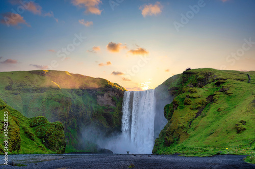 Men and women stand and raise their hands at the mighty Skogafoss Falls. In the morning  the sun rises from behind the mountains  with green grass all over the area. In southern Iceland