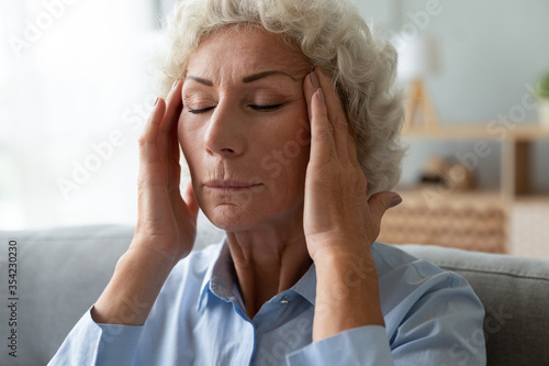 Close up face of elderly 65s woman massaging temples closed eyes reduces intense intermittent throbbing headache chronic terrible migraine, hormonal imbalance, high blood pressure hypertension concept