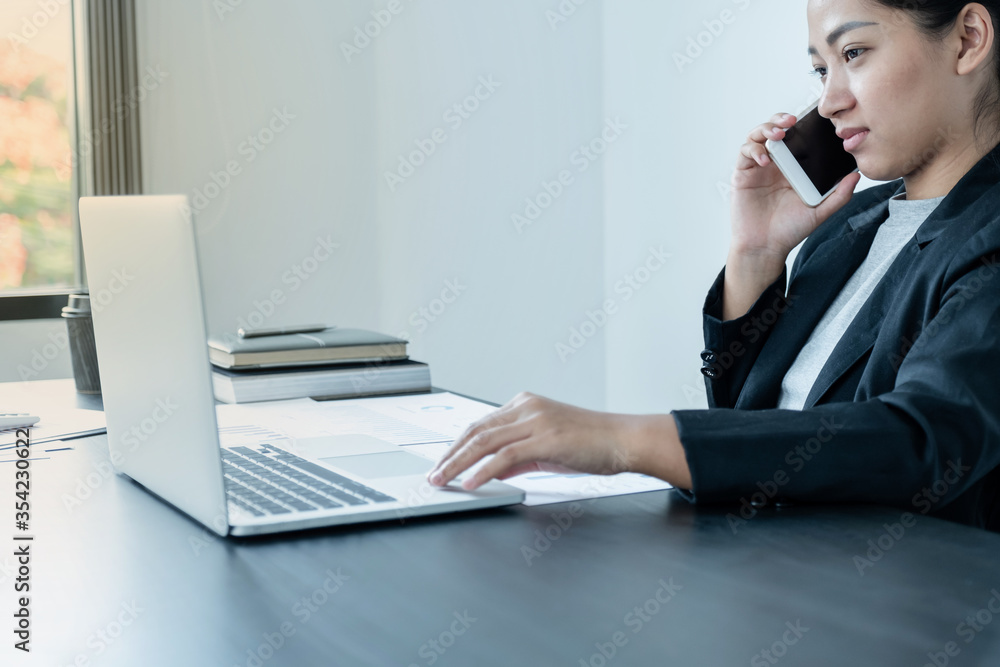 A girl businessman is talking on the phone with a smile while sitting at a desk and opening a modern working laptop in the office