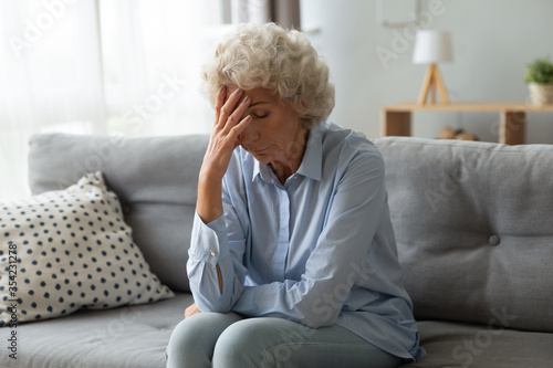 Sad elderly woman cover face with hand sit on couch lost in sad thoughts feeling miserable and lonely, dementia senile diseases, chronic migraine, unhappy unhealthy old person, life or health troubles