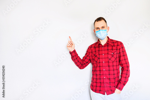 Guy in casual red plaid shirt with surgery protective mask on a face pointing finger at empty space. Isolated on light gray background