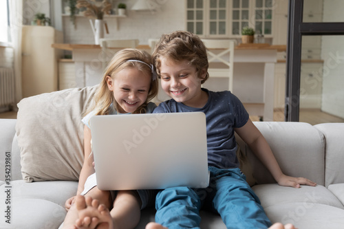 Smiling smart little boy and girl kids sit on couch in kitchen have fun using laptop together, happy small preschooler siblings brother and sister laugh watch funny videos on computer gadget at home