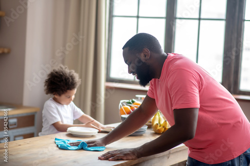 Tall dark-skinned man in a pink tshirt wiping the table while his kid helping
