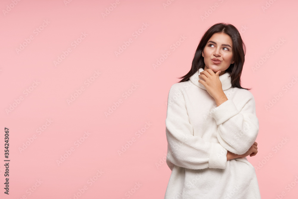 Glad young attractive brown haired female with natural makeup pursing her lips while looking pensively upwards, posing over pink background in white poloneck Glad young attractive brown haired female with natural makeup pursing her lips while looking pensively upwards, posing over pink background in white poloneck