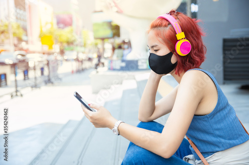 Thai Asian girl teen sitting alone for self isolation at Siam public space outdoor listening music from smartphone ware face mask, New normal lifestyle people concept.