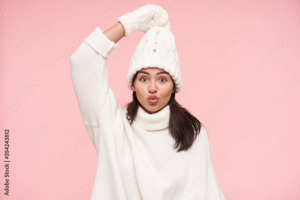 Agitated young lovely brunette female with brown loose hair pulling her hat with raised hand and looking excitedly at camera, standing over pink background Agitated young lovely brunette female with brown loose hair pulling her hat with raised hand and looking excitedly at camera, standing over pink background