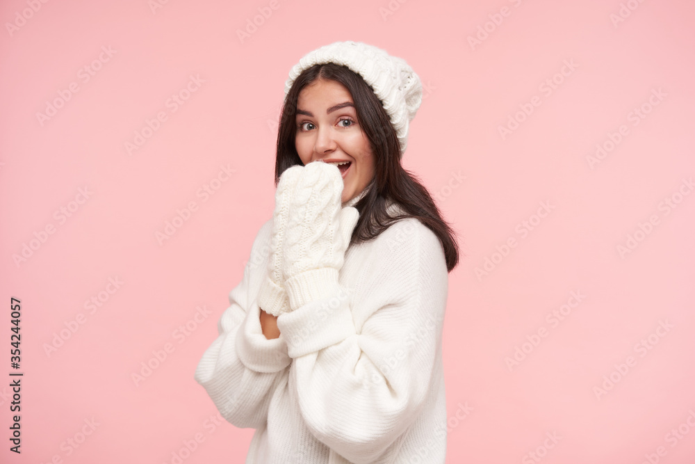 Studio photo of young agitated brunette female with natural makeup raising hands to her mouth while looking joyfully at camera, standing over pink background