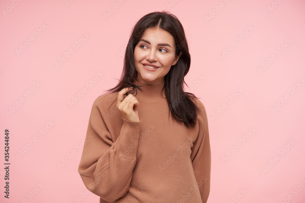 Fototapeta premium Cheerful young charming brunette female with natural makeup touching her hair with raised hand and looking gladly aside, standing over pink background