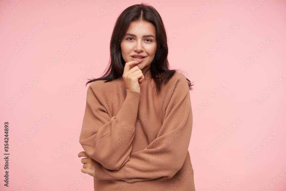 Indoor shot of young flirty pretty brunette female keeping index finger on her underlip and looking positively at camera with light smile, standing over pink background Indoor shot of young flirty pretty brunette female keeping index finger on her underlip and looking positively at camera with light smile, standing over pink background