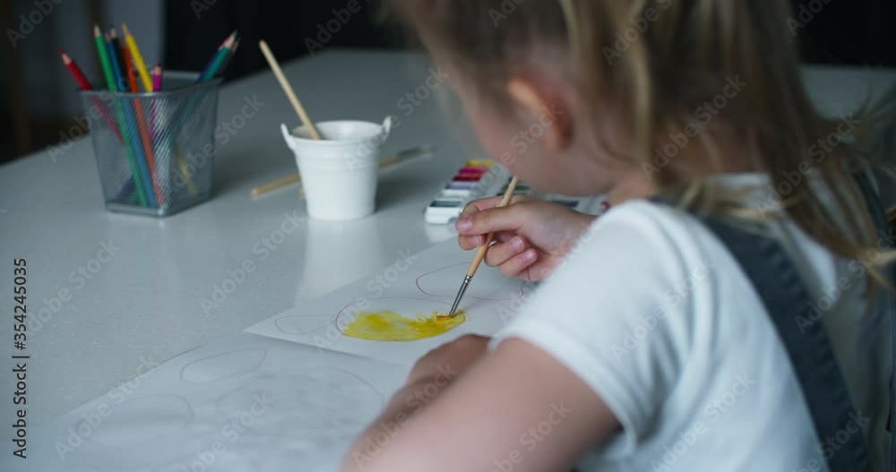 A girl child of 4 to 7 years old sits in the house and paints on paper. The child draws yellow circles with paints and pencils. A keen child is busy with his own business. Back view A girl child of 4 to 7 years old sits in the house and paints on paper. The child draws yellow circles with paints and pencils. A keen child is busy with his own business. Back view