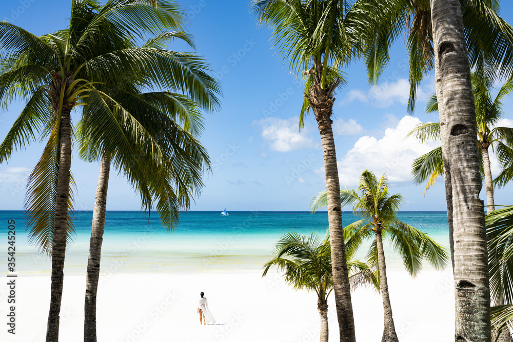 Obraz premium (Selective focus) Stunning view of woman walking on a white sand beach bathed by a turquoise sea, beautiful coconut palm trees in the foreground. White Beach, Boracay Island, Philippines.