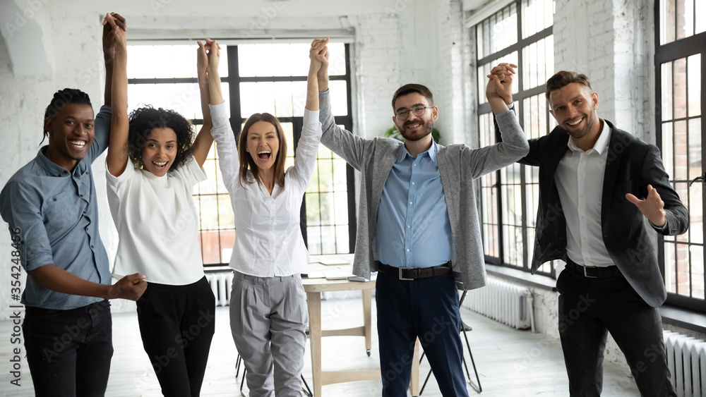 Excited diverse business people celebrating success, holding raised hands, looking at camera, smiling overjoyed employees team rejoicing achievement, laughing and screaming with joy in office room - obrazy, fototapety, plakaty 