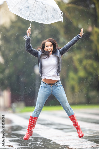 Fotografie Happy female in rubber boots and an umbrella jumps from joy on a rainy day outdo