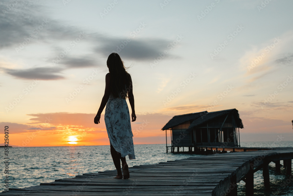 Young woman in summer dress standing on the pier with peaceful seascape enjoy view on sunset. A traveler woman  stands on a wooden pier over turquoise ocean in the Maldives and enjoys sunset