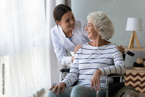 Smiling disabled elderly woman sitting in wheelchair talking with caring young nurse in living room, older generation receive homecare physical and moral support, caregiving and rehabilitation concept