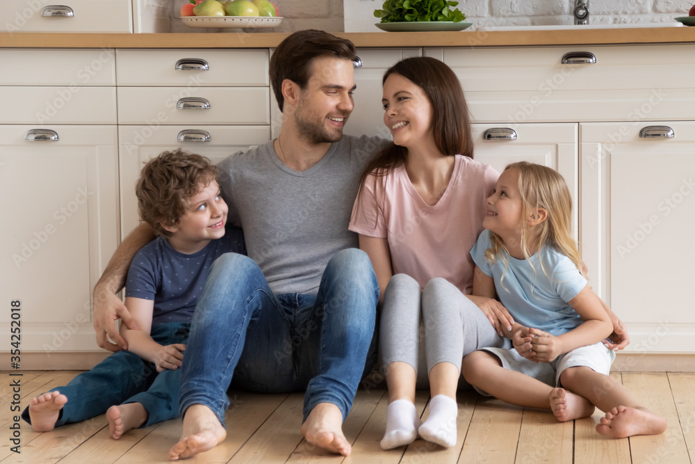 Smiling Caucasian parents with small preschooler children sit relax on warm wooden floor in modern house, happy young family with little kids rest in design renovated kitchen, relocation concept