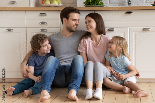 Smiling Caucasian parents with small preschooler children sit relax on warm wooden floor in modern house, happy young family with little kids rest in design renovated kitchen, relocation concept