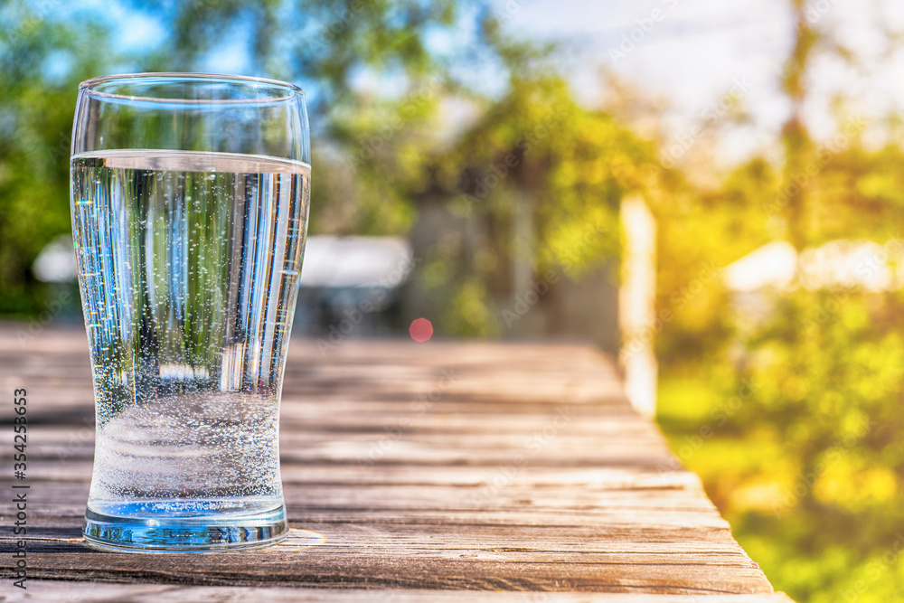 tall transparent glass with water on a wooden surface on a background of a summer landscape, blurred background tall transparent glass with water on a wooden surface on a background of a summer landscape, blurred background