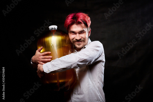 Young handsome guy with red hair in white shirt having fun with beer. Funny man with emotions on the face with large bottle of beer and black background. Brewer and barrel