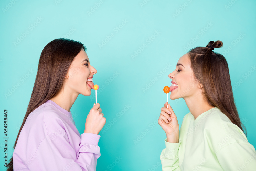 Profile photo of two funny cheerful ladies hold lollipop chupa chups hands childish mood tasty sweets wear green violet pullovers isolated pastel teal color background Profile photo of two funny cheerful ladies hold lollipop chupa chups hands childish mood tasty sweets wear green violet pullovers isolated pastel teal color background