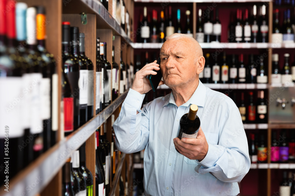 Senior man talking on mobile phone and choosing bottle of wine at the liquor store