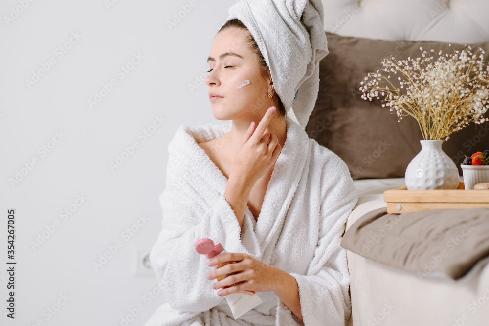 Face cream woman in bathrobe with a towel on her head after showering at home. applying skin cream on skin. Face cream woman in bathrobe with a towel on her head after showering at home. applying skin cream on skin.