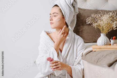 Face cream woman in bathrobe with a towel on her head after showering at home. applying skin cream on skin.