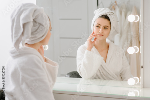 A young woman looks in the mirror in bathrobe with a towel on her head at home