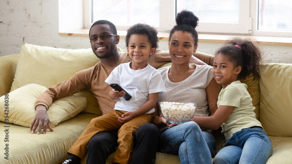 African spouses and little pretty kids spend lazy weekend together seated on couch in living room eating pop-corn choosing show or movie on TV. Happy family activity at home, free time and fun concept African spouses and little pretty kids spend lazy weekend together seated on couch in living room eating pop-corn choosing show or movie on TV. Happy family activity at home, free time and fun concept