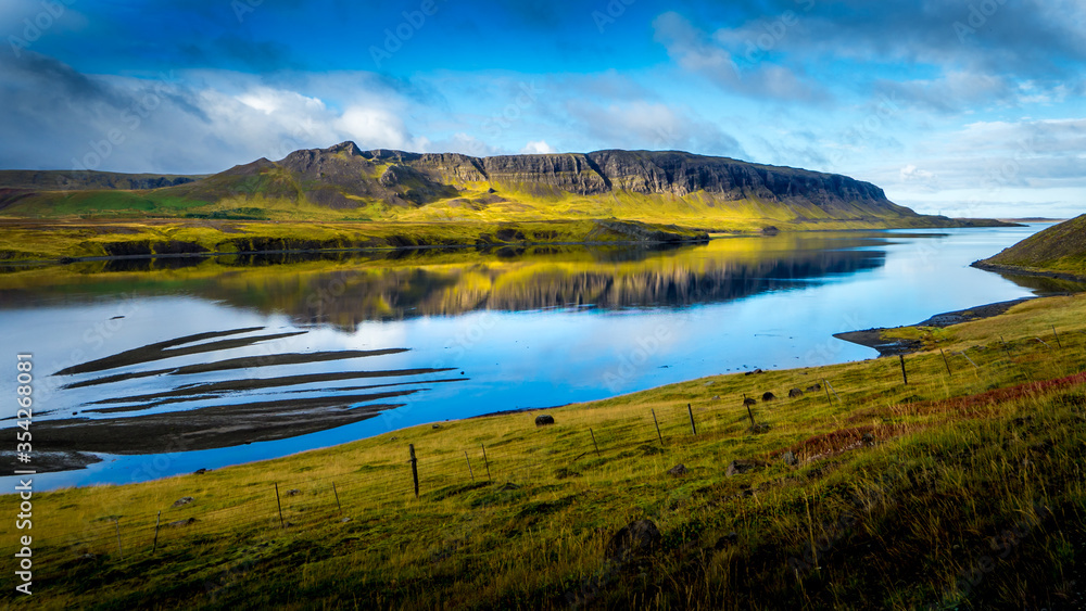 tranquil scene with view over harsh cliffs illuminated by sunshine that are reflecting in the smooth water outside at the icelandic fjord alftafjördur at snaefellsnes