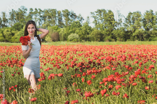 A dark, stylish, slender girl poses against the background of a poppy field with her hair flowing in the wind in the warm rays of the summer sun