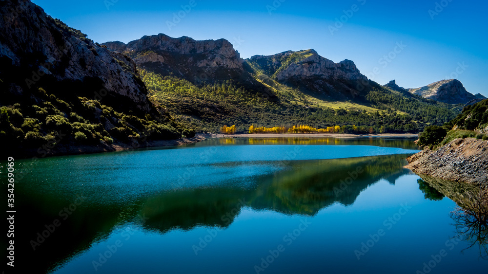 idyllic autumn view over the artificial drinking water reservoir panta del gorg blau in a valley with reflection of the mountain range serra de tramuntana and yellow colored leafs in the water