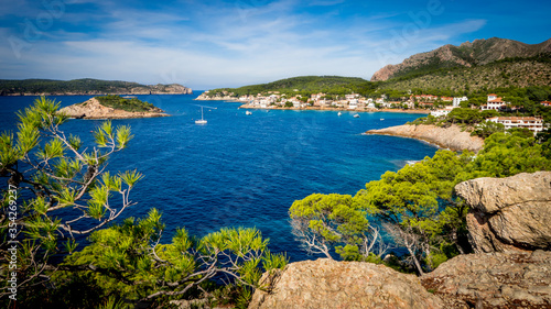picturesque sight through pine tree branches over the bay of sant elm with boats in the middle, the island sa dragonera on the left side and a mountain ridge on the right side in the background © Kris Hoobaer