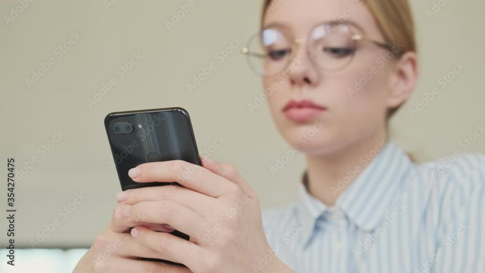 A calm focused young woman architect wearing glasses is using her smartphone while sitting in the office A calm focused young woman architect wearing glasses is using her smartphone while sitting in the office