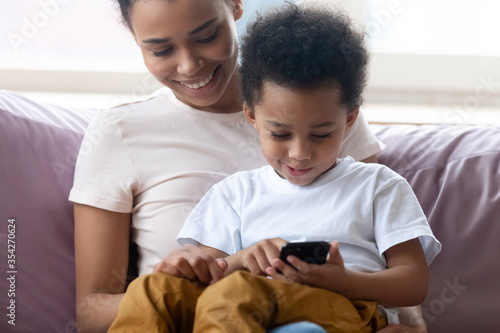 Close up view little son sit on mothers lap spend free time seated on couch holding smartphone play games, enjoy educational app on-line. Parental control, gadget overuse of younger generation concept
